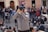 A person wearing a gray jacket and a dark cap is taking a photo with a large camera lens. They are positioned in a busy outdoor setting, surrounded by a crowd of people who are walking and gathering in groups. The background includes an old building with arched doorways.