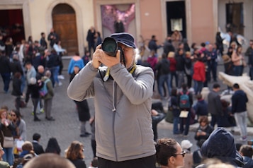 A person wearing a gray jacket and a dark cap is taking a photo with a large camera lens. They are positioned in a busy outdoor setting, surrounded by a crowd of people who are walking and gathering in groups. The background includes an old building with arched doorways.