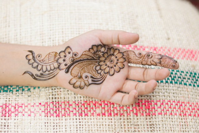 A hand is decorated with intricate henna designs featuring floral and paisley patterns. The background shows a woven mat with red and green stripes.