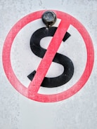 A sign displays a black letter 'S' with a red circle and diagonal line across it, indicating a prohibition. The background has a textured, weathered appearance, and a metal bolt is situated at the top of the sign.