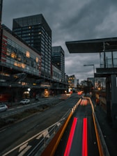 Urban scene with tall, modern buildings under a cloudy sky. The road in the foreground features streaks of light from moving vehicles, suggesting a time-lapse effect. Dimly lit street lamps and car headlights contribute to a moody atmosphere.