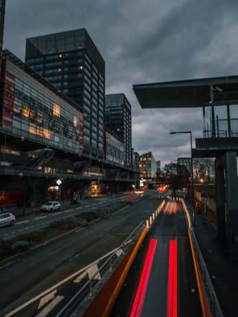 Urban scene with tall, modern buildings under a cloudy sky. The road in the foreground features streaks of light from moving vehicles, suggesting a time-lapse effect. Dimly lit street lamps and car headlights contribute to a moody atmosphere.