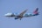 An airplane is flying across a clear blue sky. The aircraft is painted with vibrant artwork, including an image of a man with an apple in front of his face, referencing a famous surrealist painting style. The text on the plane promotes an association with Brussels, and another line mentions the Magritte Museum.