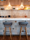 A stylish kitchen bar area featuring two modern bar stools with gray cushions and wooden legs positioned against a wall with intricate geometric tile designs. Above the countertop are two pendant lights with wooden covers that emit a warm glow. The countertop displays small potted plants and a range of kitchenware, while the wall behind is covered with beige tiles.