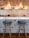 A stylish kitchen bar area featuring two modern bar stools with gray cushions and wooden legs positioned against a wall with intricate geometric tile designs. Above the countertop are two pendant lights with wooden covers that emit a warm glow. The countertop displays small potted plants and a range of kitchenware, while the wall behind is covered with beige tiles.