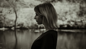 A black and white profile of a person with short hair standing in front of a blurred outdoor background, possibly near a body of water and trees.
