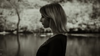 A black and white profile of a person with short hair standing in front of a blurred outdoor background, possibly near a body of water and trees.