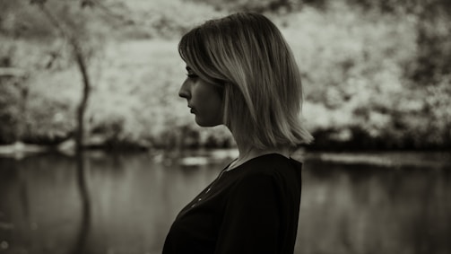 A black and white profile of a person with short hair standing in front of a blurred outdoor background, possibly near a body of water and trees.