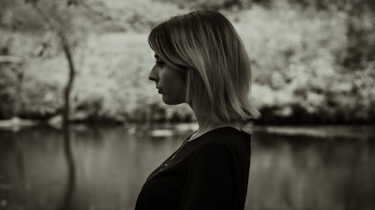 A black and white profile of a person with short hair standing in front of a blurred outdoor background, possibly near a body of water and trees.