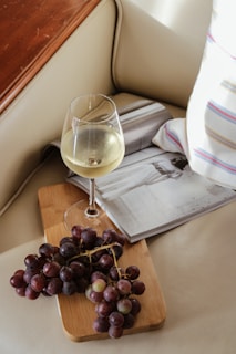 A wine glass filled with white wine sits on a wooden cutting board, which also holds a bunch of purple grapes. An open magazine is placed nearby on a cream-colored cushioned surface, with a striped pillow partially visible on the side. The setting suggests a relaxed and cozy atmosphere.