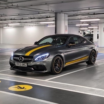 A sleek, modern Mercedes-Benz with a sports design is parked in a well-lit underground parking garage. The car features distinctive yellow racing stripes running along its body, contrasting with its metallic gray color. Ambient lighting highlights the car's elegant curves and sophisticated design.