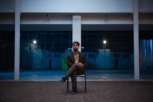 A man sits alone on a chair in an industrial, dimly lit setting. The space is large and open with high ceilings, featuring structural beams and a dark blue background. The man appears thoughtful or introspective, with subtle illumination highlighting his presence.