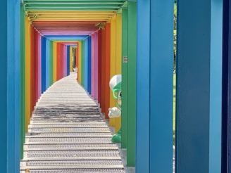 A colorful walkway with a series of rainbow-colored beams forming a tunnel effect. Each beam is painted in a different vibrant color, creating a gradient spectrum. The path is made of metal grating and stretches into the distance, adding to the sense of depth. On the right, behind one of the beams, a green cartoon character with goggles and a white hat peeks out playfully.