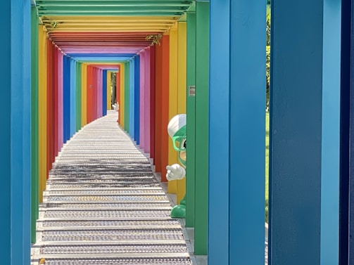 A colorful walkway with a series of rainbow-colored beams forming a tunnel effect. Each beam is painted in a different vibrant color, creating a gradient spectrum. The path is made of metal grating and stretches into the distance, adding to the sense of depth. On the right, behind one of the beams, a green cartoon character with goggles and a white hat peeks out playfully.