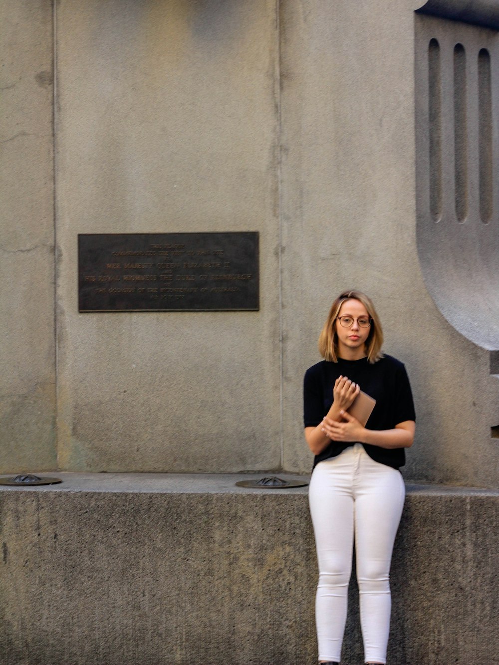 woman in black long sleeve shirt and white pants leaning on gray concrete wall