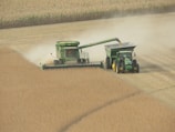 A green combine harvester is actively harvesting grain in a large field. Dust clouds are forming behind it, indicating movement and activity. Accompanying the harvester is a tractor and a grain cart positioned beside it, collecting the harvested crop.