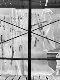 A plastic sheet with visible tape repairs is spread over a window, divided by a central mullion. The exterior reveals a brick wall, with rough wooden planks at the base of the window and a partly visible architectural structure above.