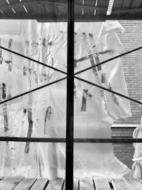 A plastic sheet with visible tape repairs is spread over a window, divided by a central mullion. The exterior reveals a brick wall, with rough wooden planks at the base of the window and a partly visible architectural structure above.