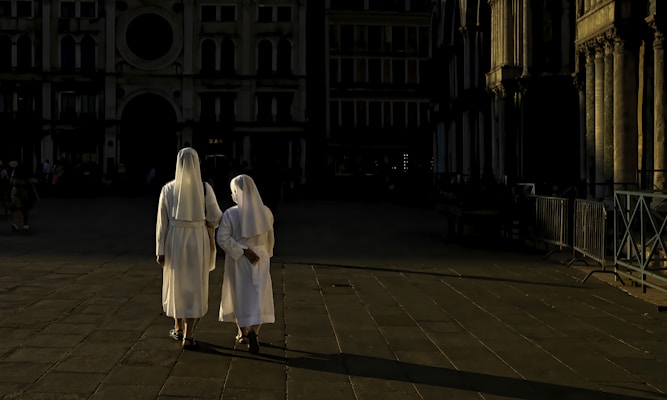 Two nuns dressed in white habits are walking away on a paved, sunlit path next to a historic building with ornate columns. The background is cast in shadow, providing a stark contrast to the illuminated areas. The scene evokes a sense of calm and reflection as the figures walk side-by-side.
