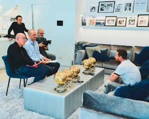 A group of people are gathered in a modern living room. One person is seated on one side near a coffee table, which displays multiple golden trophies. Three individuals are seated facing this person, two with papers, and another stands in the back with a camera. The room is decorated with a plush gray sofa and blue cushions, and family photos are displayed on the wall shelves.