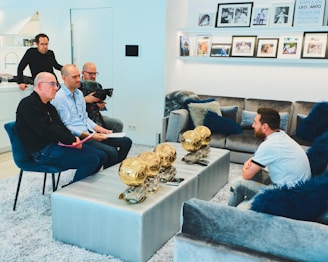 A group of people are gathered in a modern living room. One person is seated on one side near a coffee table, which displays multiple golden trophies. Three individuals are seated facing this person, two with papers, and another stands in the back with a camera. The room is decorated with a plush gray sofa and blue cushions, and family photos are displayed on the wall shelves.