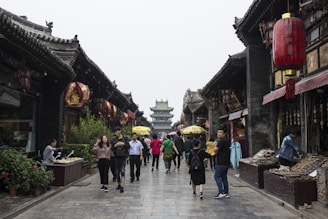 A bustling ancient street with traditional architecture, featuring ornate wooden buildings adorned with red lanterns. People are walking and shopping along the stone-paved path, contributing to a lively atmosphere. Stalls on either side display various goods, while the background features a pagoda.