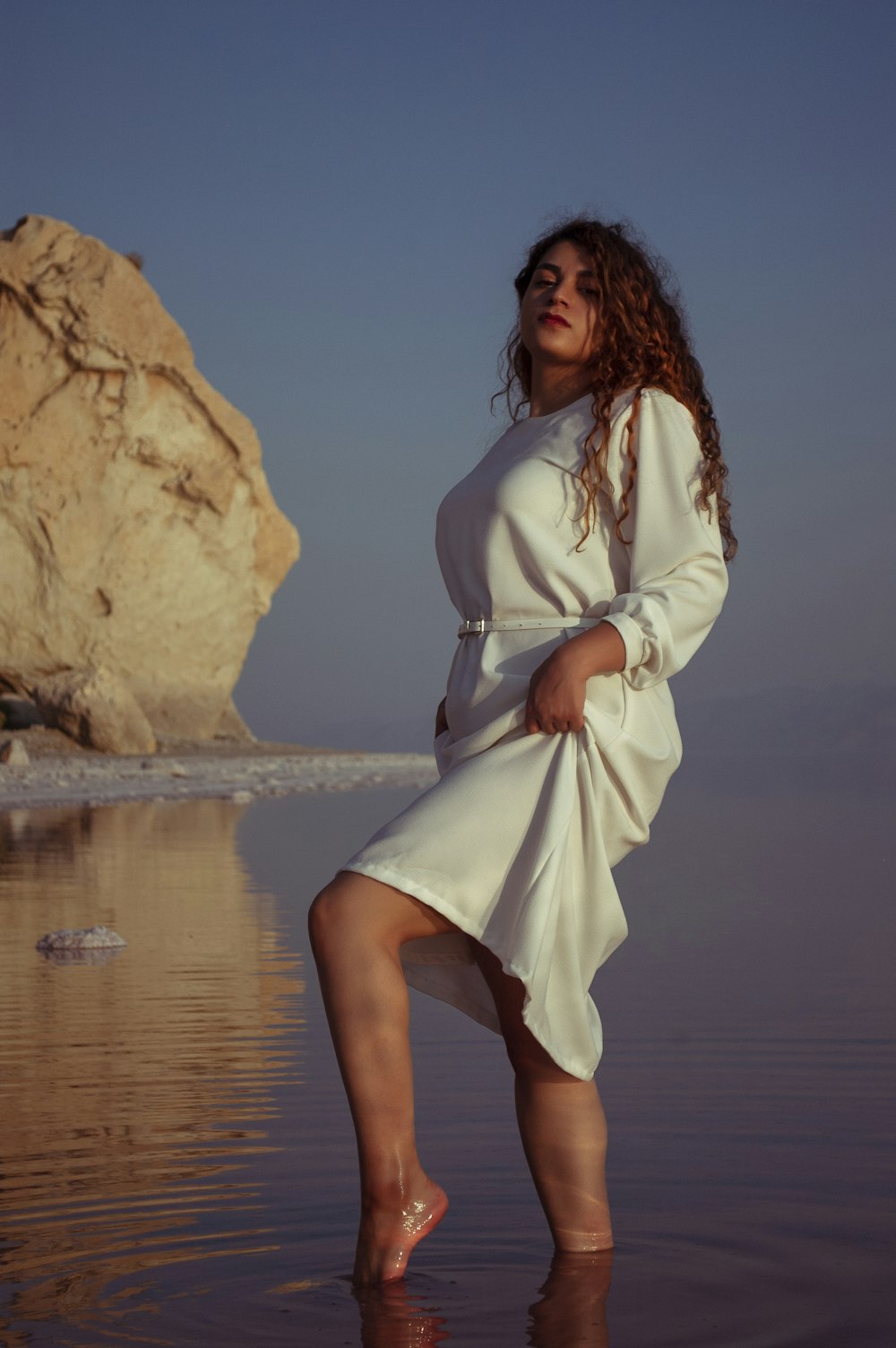 woman in white long sleeve dress standing on brown rock near body of water during daytime