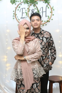 A couple standing in front of a decorative backdrop adorned with greenery and circular frames containing the letters 'E' and 'H'. The woman on the left is wearing a hijab and a patterned dress, covering one eye with her hand that displays a ring, while the man beside her wears a matching patterned shirt. A wooden stool is visible beside them.