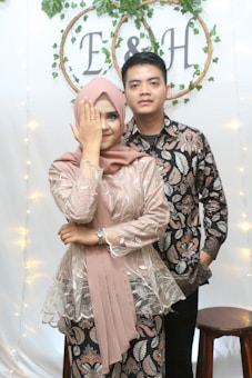 A couple standing in front of a decorative backdrop adorned with greenery and circular frames containing the letters 'E' and 'H'. The woman on the left is wearing a hijab and a patterned dress, covering one eye with her hand that displays a ring, while the man beside her wears a matching patterned shirt. A wooden stool is visible beside them.