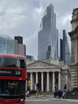 A cityscape featuring a mix of modern and classical architecture. A red double-decker bus with the signage 'Waterloo 26' is prominent in the foreground. Behind the bus is a grand neoclassical building with columns and detailed sculptures on its pediment. In the background, several modern skyscrapers, including one with a distinctive angular design, rise into the cloudy sky. People can be seen walking on the sidewalk.