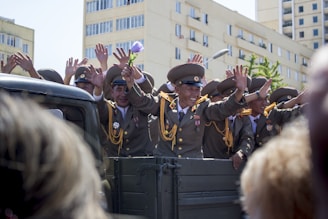 A group of uniformed military officers is seen standing on a vehicle, cheering and waving to a crowd. The officers are dressed in formal military attire, adorned with medals and gold insignias. One officer is holding a flower, and they are surrounded by modern apartment buildings and trees, suggesting an urban setting.