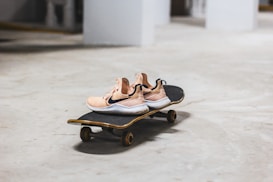 A pair of beige sneakers with black swoosh logos placed on a skateboard on a smooth concrete floor. The skateboard has a black grip tape and appears unused. The setting resembles an indoor area with white columns and a minimalistic, slightly industrial look.