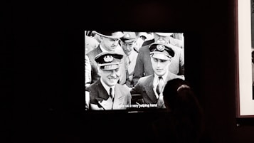 An old black and white photograph displayed on a screen shows a group of uniformed individuals, likely from a historical military or aviation context. The subtitles at the bottom read 'gave us a very helping hand.' One person in the foreground stands partially silhouetted, viewing the image.