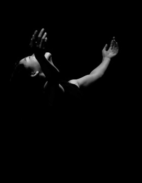 A dramatic black and white image captures the side profile of a person with arms reaching upward, their face partially illuminated against a dark background. The light accentuates the expression and posture, conveying a sense of emotion.