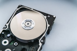 A close-up view of an opened hard disk drive, showcasing the shiny, reflective surface of the platter and the read/write head assembly. The metallic components are contrasted against a neutral background.