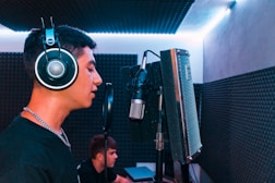 A person wearing large headphones is singing or speaking into a microphone in a music recording studio. The room features soundproofing foam on the walls and dim blue lighting, adding to the focused atmosphere.