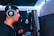 A person wearing large headphones is singing or speaking into a microphone in a music recording studio. The room features soundproofing foam on the walls and dim blue lighting, adding to the focused atmosphere.