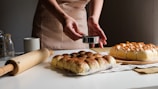 Hands sprinkle a spice or powder over baked goods, which are neatly arranged on a table. The scene includes a rolling pin, a mug, and jars, suggesting a baking or kitchen setting.