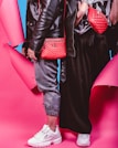Two individuals dressed in trendy streetwear fashion stand side by side, featuring black leather jackets and red bags against a vibrant pink and blue background. One person wears gray joggers and white sneakers, while the other wears a black skirt and white sneakers. The red bags are prominently displayed with quilted designs.