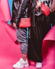 Two individuals dressed in trendy streetwear fashion stand side by side, featuring black leather jackets and red bags against a vibrant pink and blue background. One person wears gray joggers and white sneakers, while the other wears a black skirt and white sneakers. The red bags are prominently displayed with quilted designs.