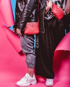 Two individuals dressed in trendy streetwear fashion stand side by side, featuring black leather jackets and red bags against a vibrant pink and blue background. One person wears gray joggers and white sneakers, while the other wears a black skirt and white sneakers. The red bags are prominently displayed with quilted designs.
