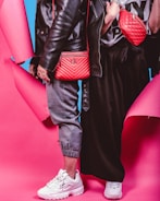Two individuals dressed in trendy streetwear fashion stand side by side, featuring black leather jackets and red bags against a vibrant pink and blue background. One person wears gray joggers and white sneakers, while the other wears a black skirt and white sneakers. The red bags are prominently displayed with quilted designs.