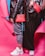 Two individuals dressed in trendy streetwear fashion stand side by side, featuring black leather jackets and red bags against a vibrant pink and blue background. One person wears gray joggers and white sneakers, while the other wears a black skirt and white sneakers. The red bags are prominently displayed with quilted designs.