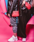 Two individuals dressed in trendy streetwear fashion stand side by side, featuring black leather jackets and red bags against a vibrant pink and blue background. One person wears gray joggers and white sneakers, while the other wears a black skirt and white sneakers. The red bags are prominently displayed with quilted designs.