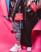 Two individuals dressed in trendy streetwear fashion stand side by side, featuring black leather jackets and red bags against a vibrant pink and blue background. One person wears gray joggers and white sneakers, while the other wears a black skirt and white sneakers. The red bags are prominently displayed with quilted designs.