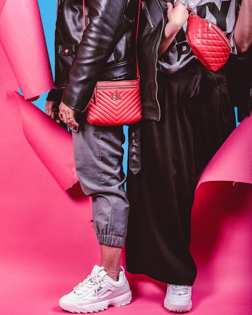 Two individuals dressed in trendy streetwear fashion stand side by side, featuring black leather jackets and red bags against a vibrant pink and blue background. One person wears gray joggers and white sneakers, while the other wears a black skirt and white sneakers. The red bags are prominently displayed with quilted designs.