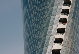 A modern skyscraper features a curved glass facade with a pattern of diagonal lines. Balconies with glass railings are visible along one side of the building.