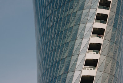 A modern skyscraper features a curved glass facade with a pattern of diagonal lines. Balconies with glass railings are visible along one side of the building.
