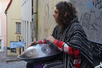 A person is sitting on a bench, playing a handpan in an urban setting. The walls around are covered with various graffiti. The person is wearing a black and white patterned poncho over a red checkered shirt. A narrow cobblestone alleyway is visible, lined with pastel-colored buildings.