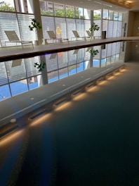 A calm indoor swimming pool area with clear water reflecting the surroundings. On one side, there are several white lounge chairs and potted plants arranged neatly. Large windows allow natural light to flood the space, and a horizontal reflection creates a symmetry in the pool.
